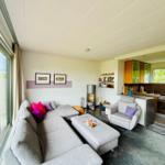 Wohnzimmer mit Sofa, Sessel und Küchenbereich. Große Fenster und Holzelemente.