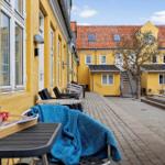 Outdoor area with tables, chairs, and blue blanket. Yellow buildings with red roof and pool in background.
