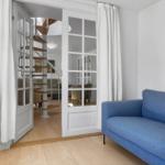 Ein Raum mit blauem Sofa, weißen Vorhängen und einer Tür zum Treppenhaus.