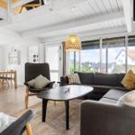 Wohnzimmer mit Sofa, Sessel und Kamin. Durchgehende Fensterfront mit Blick auf Außenbereich.