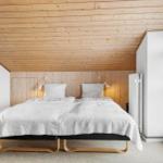 Schlafzimmer mit Doppelbett, Holzdecke und Fenster. Wandlampe und Heizung sichtbar.