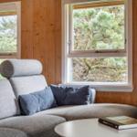 Graue Couch mit Kissen vor Holzwänden und Fenstern mit Blick auf Bäume.