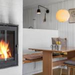 Kamin mit Flammen, Esstisch mit Holzbank und Stühlen, Wand mit Bildern und Pendelleuchte.
