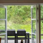 Sliding glass doors open to a terrace with table and chairs. Green garden in the background.