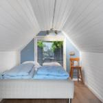 Schlafzimmer mit Bett, Stuhl und Fenster. Wände und Decke sind weiß verkleidet.