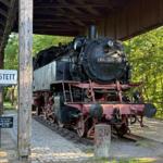 Dampflokomotive 064-355-1 steht auf Bahnsteig unter Dach. Schild zeigt Hillstett. Grün hinter dem Bahnhof.