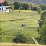 Golfplatz mit Golfwagen und Spieler. Im Hintergrund moderne Villa und Wald.