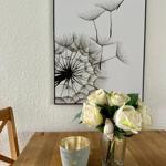 Holztisch mit Blumen in Glas und Kerze vor Wandbild mit Löwenzahn.