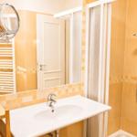 Badezimmer mit Dusche, Waschbecken und Spiegel. Holzregal und Heizung sichtbar.