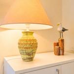 Tischlampe mit gelbem Schirm auf weißen Schrank. Holzdekoration mit Vogelfigur daneben.