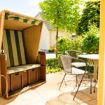 Terrasse mit Strandkorb, Tisch und zwei Stühlen. Grüner Garten im Hintergrund.