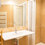 Bad mit Dusche, Waschbecken und Spiegel. Holzregal und Heizkörper sichtbar.