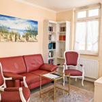 Ein Wohnzimmer mit rotem Sofa, weißen Stühlen und Bücherregal. An der Wand hängt ein Strandbild.