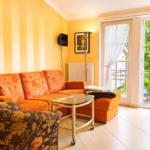 Wohnzimmer mit orangefarbenem Sofa, Glas-Tisch und Balkontür zu grünem Außenbereich.