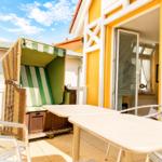 Terrasse mit Strandkorb, Tischen und Stühlen vor gelbem Haus mit Schiebetür.