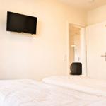 Schlafzimmer mit Bett, Wand-TV und Kleiderschrank. Tür führt in angrenzenden Raum.