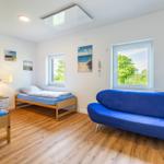 Zimmer mit zwei Betten, blauem Sofa und Holzboden. Fenster zeigen grüne Bäume.