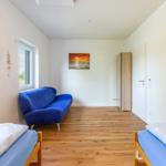 Zimmer mit zwei Betten, blauem Sofa und Holzboden. Wandbilder zeigen Meereslandschaften.