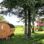 Holzsauna und Kinderbereich unter Bäumen im Gras.