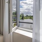 Durch das Fenster blickt man auf einen Kanal mit grünen Ufern und Häusern im Hintergrund.