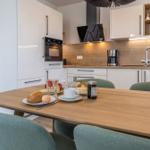 Essbereich mit Holztisch und grünen Stühlen. Küche mit weißen Schränken und Holzrückwand.