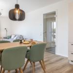 Essbereich mit Holztisch, grünen Stühlen und Küchenzeile. Im Hintergrund Wohnbereich mit Sofa.