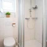 Badezimmer mit Dusche, Toilette und Fenster. Weiße Fliesen, Handtuchhalter und Pflanze.
