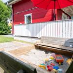 Holzspielplatz mit Sand und bunten Spielzeugen vor rotem Haus mit weißem Zaun.