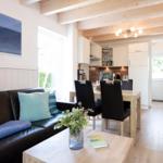 Wohnzimmer mit Sofa, Esstisch und Küche im Hintergrund. Holzbalken und Wandverkleidung.