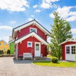 Rotes Ferienhaus mit weißer Tür und Bank vorne. Grasfläche und Kiesweg. Nebenhaus mit Gitterzaun.