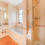 Bad mit Dusche, Badewanne und Waschbecken. Fenster, Spiegel und Bodenfliesen.