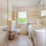 Badezimmer mit Dusche, Toilette, Waschmaschine und Fenster mit Blick auf Grün.