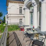 Terrasse mit Tisch, Stühlen und Flasche. Blick auf Straße und Grünflächen.