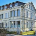 Villa Sandrose mit hellen Fassaden und Säulen. Vorderhof mit Rasen und Gehweg.