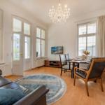 Wohn- und Essbereich mit Sofa, Esstisch und Kronleuchter. Holzboden und große Fenster.