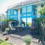 Terrasse mit Sitzgelegenheiten und Pflanzen vor einem Haus mit blauen Fenstern.