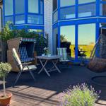 Terrasse mit Sitzgelegenheiten und Pflanzen vor einem Haus mit blauen Fensterrahmen.