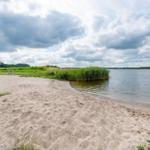 Strand mit Sand, Gras und Wasser unter bewölktem Himmel.