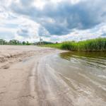 Strand mit Sand, Wasser und Gras. Im Hintergrund sind Bäume und ein Fußballtor zu sehen.