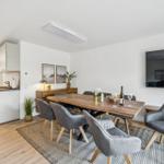 Essbereich mit Holztisch, grauen Stühlen und Fernseher an der Wand.
