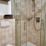 Dusche mit Glaswand und Duschbrause, Wandfliesen und Toilette sichtbar.