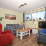 Wohnzimmer mit Sofa, zwei Sesseln, Couchtisch und TV. Fenster zeigt Leuchtturm.