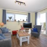 Wohnzimmer mit Sofa, Sessel und Fernseher. Fenster und Glastür führen auf eine Terrasse.