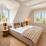Schlafzimmer mit Doppelbett, zwei Fenstern und goldener Sitzgelegenheit.