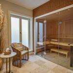 Sauna mit Glaswand und Holzbank. Nebenraum mit Sitzgelegenheit und Pflanze.