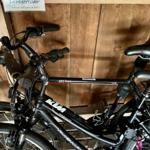 Zwei schwarze Fahrräder stehen in einer Holzwandgarage. Ein Schild zeigt Leihfahrräder an.