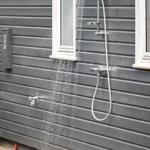 Outdoor shower on gray wooden house wall with water stream and plant in foreground.