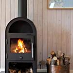 Wood stove with burning logs, beside bucket with firewood against wood wall.