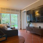 Living room with sofa, TV, and terrace door. Terrace with green area visible.