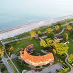 Haus mit rotem Dach liegt am Strand. Grünflächen und Bäume umgeben die Anlage.